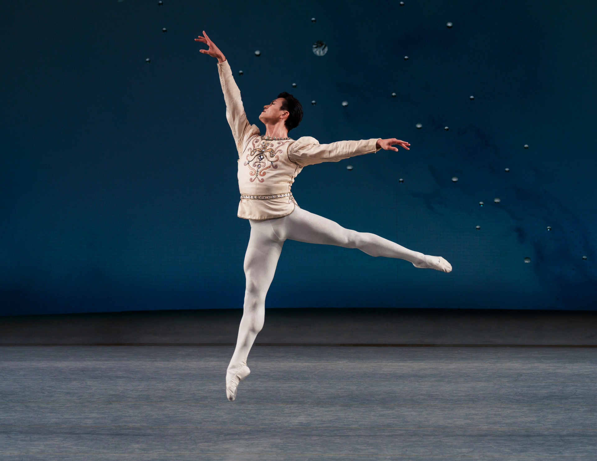 Diamonds series — Diamonds — soaring grand jeté in cream tunic against deep blue backdrop